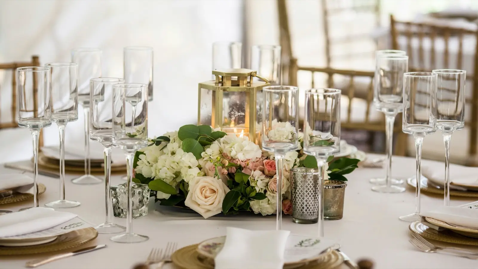 A laid wedding table with glasses, a gold lantern and a low centerpiece of roses and hydrangea