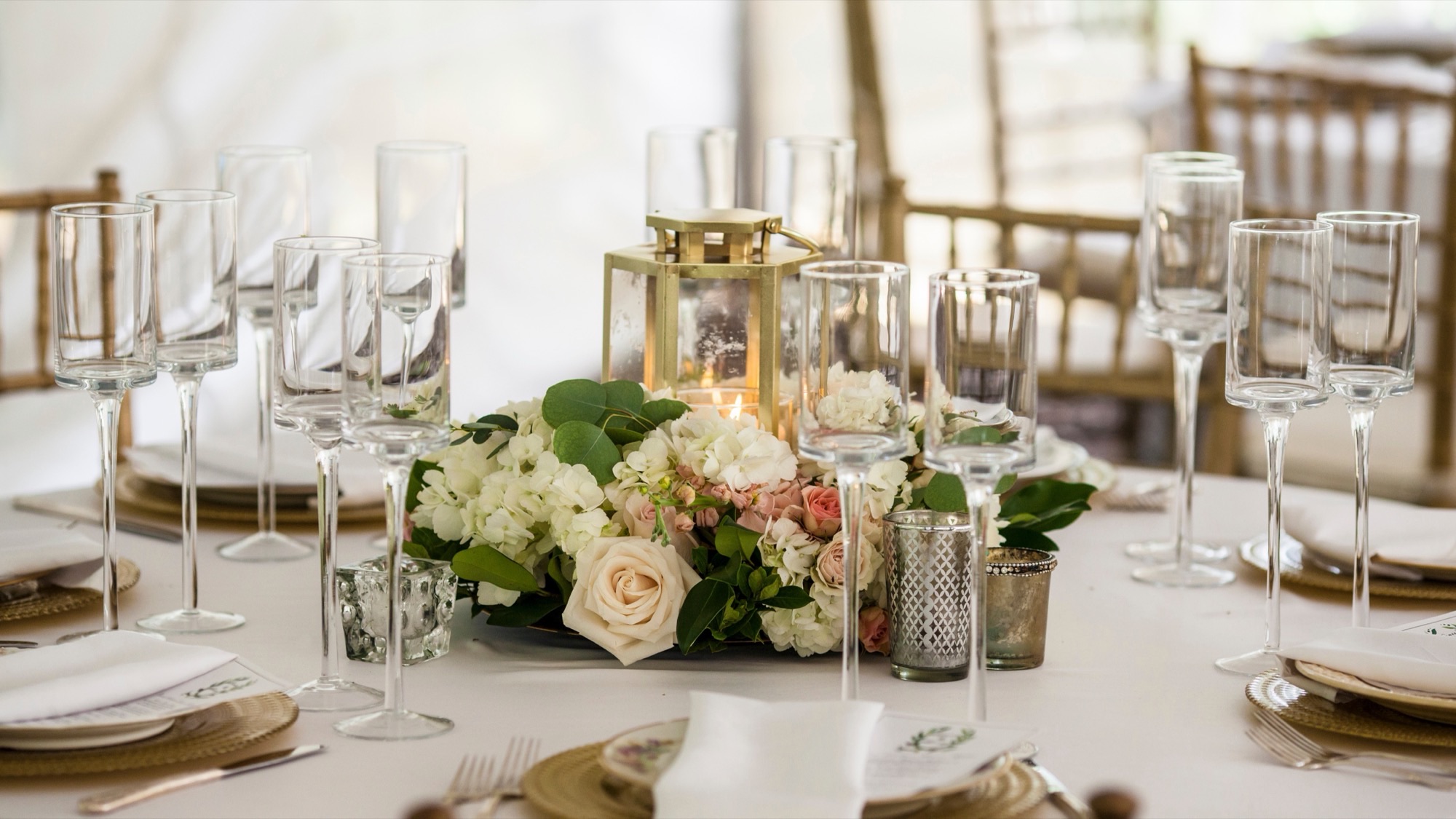 A laid wedding table with glasses, a gold lantern and a low centerpiece of roses and hydrangea